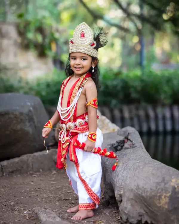 A full-length portrait of a young boy dressed as Lord Krishna, holding a flute and posing confidently in a park.
