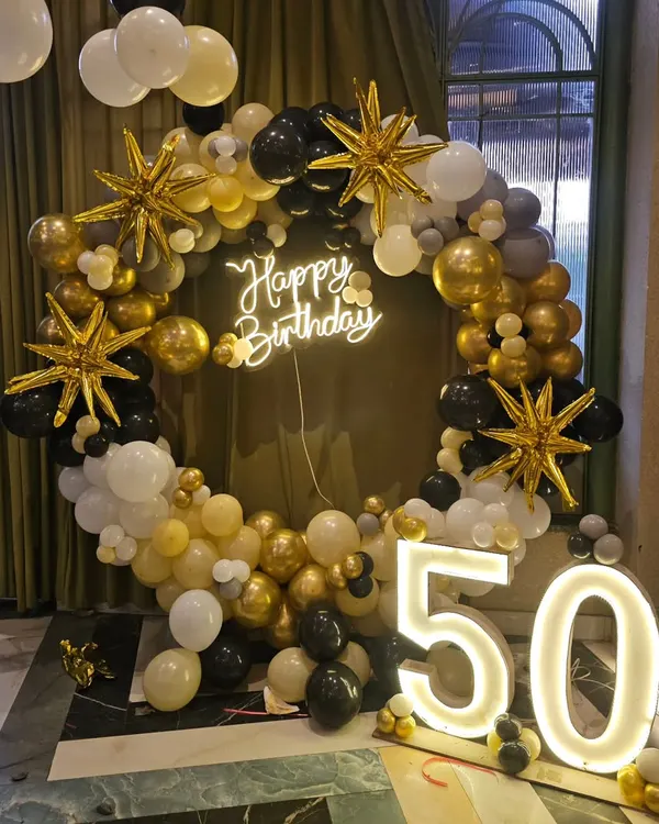 An elegant 50th birthday decoration featuring a black, gold, and white balloon ring, a 'Happy Birthday' neon sign, and large light-up '50' numbers.