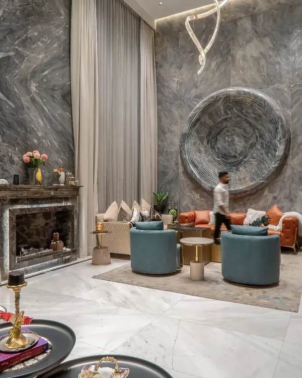 A view of the double-height living room, showcasing the grand scale and material richness. The design features a marble-clad fireplace, elegant drapery, and a curated selection of furniture that balances comfort with sophisticated style.
