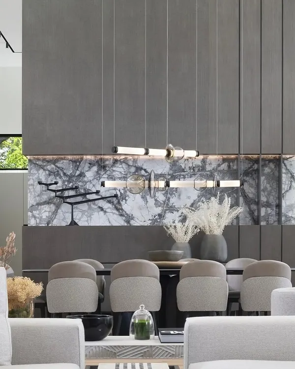 A horizontal Luna series chandelier creates a dramatic focal point above a modern dining area. The composition of illuminated tubes and hand-blown glass elements is set against a sophisticated backdrop of dark marble and panelled walls, showcasing how I integrate grand lighting statements into luxurious living spaces.