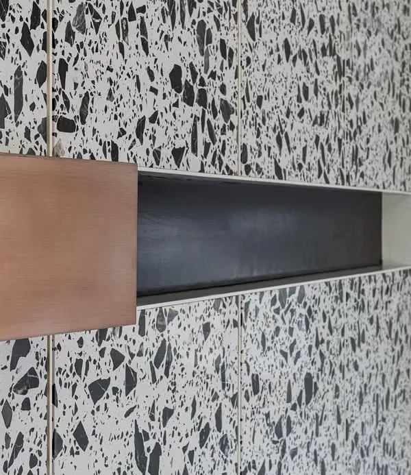 A close-up of the copper cuboid wedged into the terrazzo paneling in 'A Wedged Haus'. The reflective nature of the polished copper contrasts sharply with the matte, composite texture of the terrazzo stone.