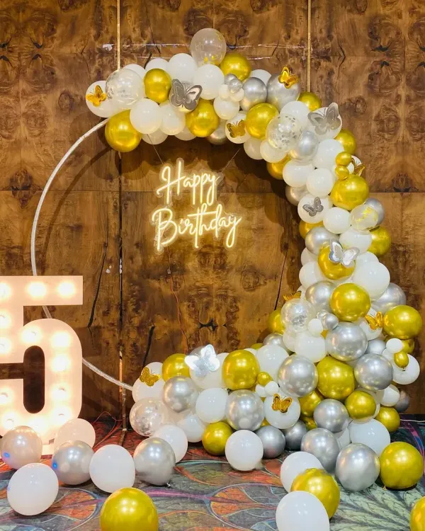 A modern and chic birthday setup featuring a large ring frame wrapped in a crescent of gold, white, and silver balloons. The design is completed with a "Happy Birthday" neon sign, a large light-up number '5', and delicate butterfly accents for a touch of magic.