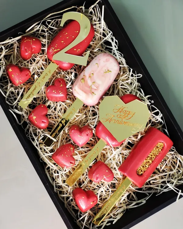 This anniversary gift box is designed for a truly personal celebration. It includes custom-engraved cakesicles and heart-shaped chocolates, all elegantly arranged on a bed of shredded paper.