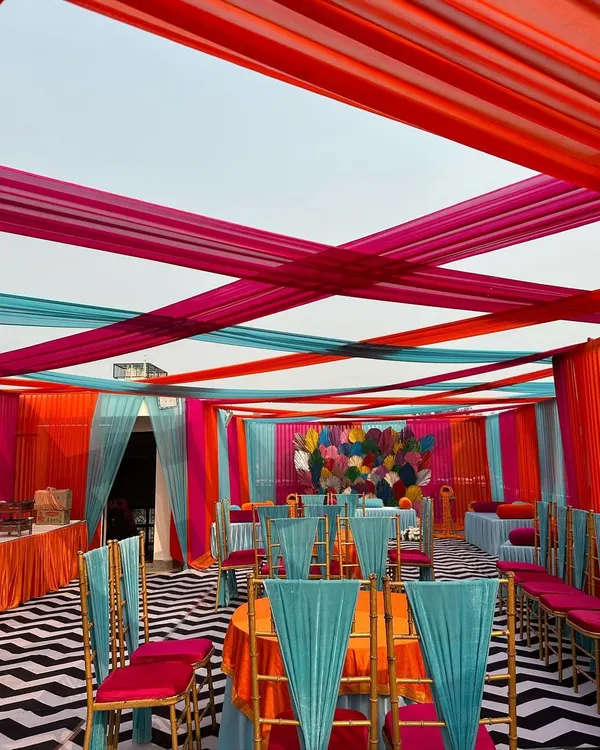 A stunningly vibrant Mehendi setup with a canopy of pink, orange, and turquoise drapes. The chevron-patterned floor and colorful seating created a lively and festive atmosphere.