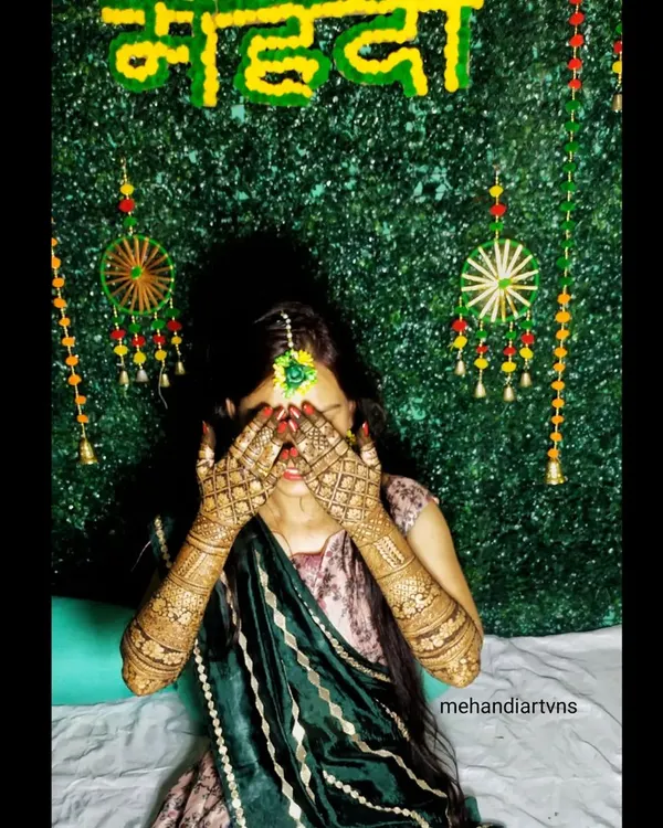 A classic shy bride pose behind her beautifully decorated hands. The green background makes the rich color of the mehandi stand out.