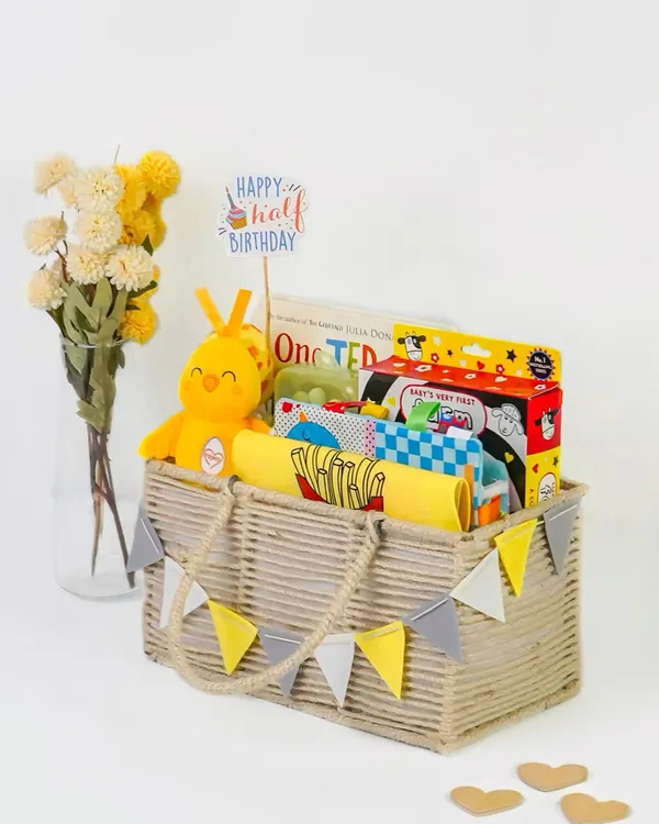 A 'Happy Half Birthday' themed baby hamper, beautifully arranged in a rustic basket. The bright yellow and grey color scheme is modern and cheerful.