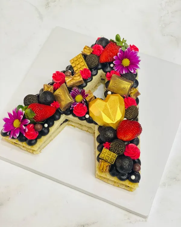 A stunning monogram cake in the shape of the letter 'A'. This one is loaded with fresh strawberries, blueberries, Lindt chocolates, and a geometric chocolate heart.