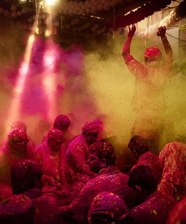 Inside a temple during Barsana Holi, where clouds of pink and yellow gulal fill the air. A single ray of light illuminates the scene of joyful celebration.