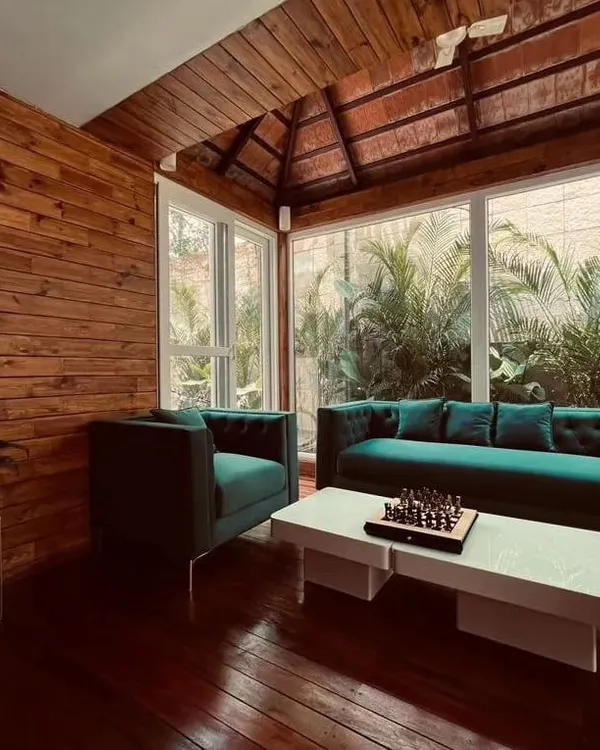 An interior view of a stunning patio room at Zonasha Paradiso for Mr. Amar Nagaram, founder of Virgio. This space features rich Brazilian Ipe wood flooring, wood-paneled walls, and a Mangalore tile roof, furnished with elegant teal sofas for a luxurious feel.