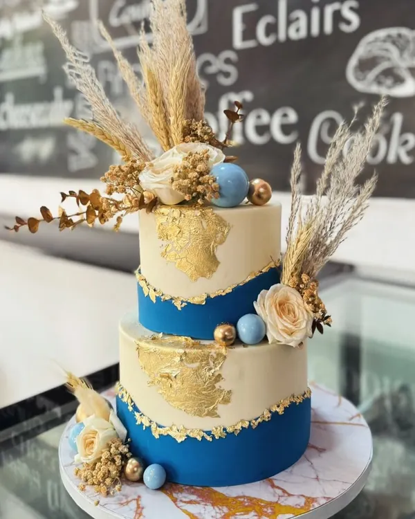 A stunning two-tiered eggless cake perfect for an elegant celebration like an engagement or a milestone anniversary. This design features a bold royal blue base, delicate gold leaf accents, and is beautifully finished with dried pampas grass and cream-colored roses.
