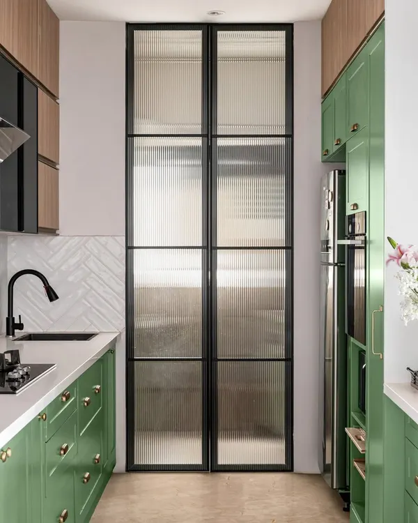A full view of our Elle Decor-featured kitchen. This galley layout includes a utility area separated by a fluted glass door, built-in appliances, and a beautiful contrast between the green cabinets and wood-tone overheads.