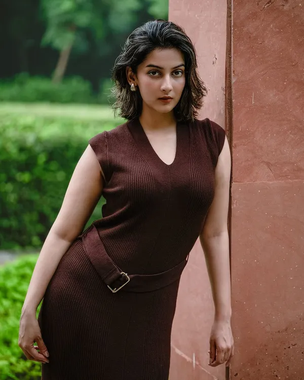 A powerful outdoor portrait of a model in a form-fitting brown dress, leaning against a wall. The pose and direct gaze create a confident and striking image.