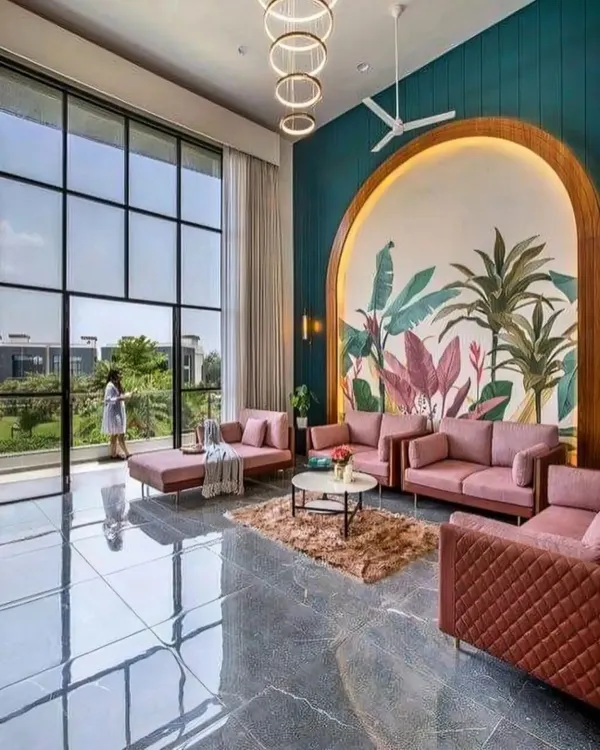 This expansive living room in a luxury villa features double-height windows that flood the space with natural light. We used a soft color palette with blush pink sofas and a statement tropical mural within an arched niche to create a sophisticated yet welcoming atmosphere.
