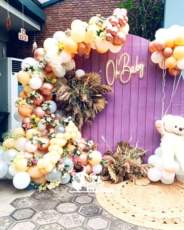 This rustic-themed baby shower featured a beautiful photo backdrop with a purple wooden panel, an 'Oh Baby' neon sign, and a large teddy bear floating on a cloud of balloons. The look was softened with pampas grass and a jute rug.