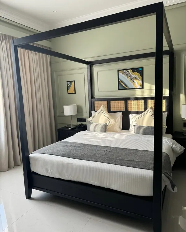 A modern four-poster canopy bed with a clean, minimalist frame in a black finish. The headboard features teak and cane panels, combining modern and traditional elements. This was part of a hotel project.