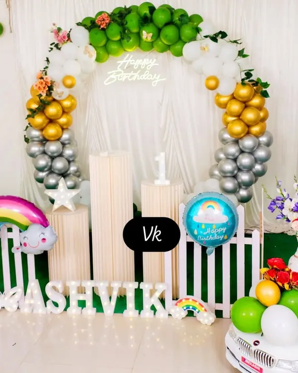 This first birthday setup features a circular balloon arch in green, gold, and silver. It includes a neon sign, marquee name lighting, and fun props like a rainbow balloon and a mini car.
