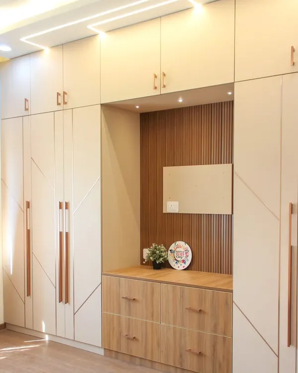 A full view of a spacious bedroom wardrobe in beige laminate with geometric routing and long, rose gold handles. The design includes a built-in dresser nook with a wood-paneled back and drawers.