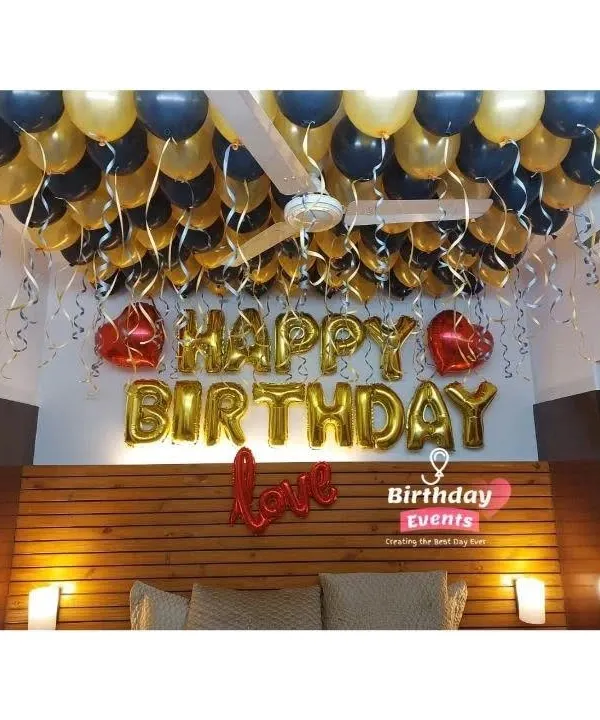 A classic bedroom birthday surprise. The ceiling is covered with black and gold balloons with dangling ribbons, complemented by gold foil "Happy Birthday" and heart-shaped balloons.
