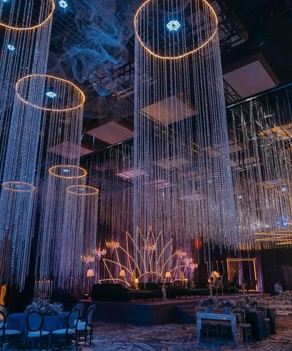 A wide view of the celestial-themed sangeet, where pillars of cascading crystals and illuminated rings hang from the ceiling. The design creates a magical, starry ambiance, with a neon lotus stage as the focal point for the evening's performances.