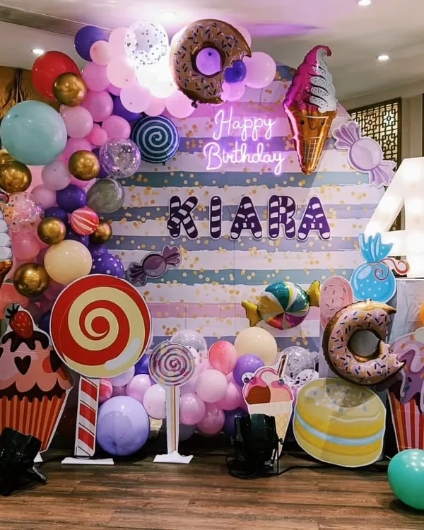 A detailed look at a Candyland birthday theme, complete with a striped backdrop, neon "Happy Birthday" sign, and various props like giant cupcakes, lollipops, and donuts.