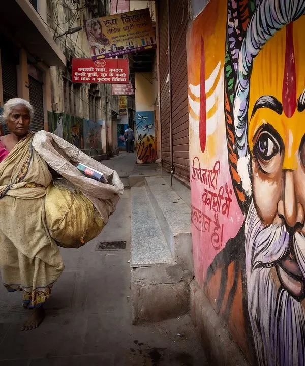 An elderly woman walks through a narrow lane in Varanasi, passing a vibrant mural of a sadhu. This image captures the intersection of daily life, art, and spirituality in the city's ancient streets.