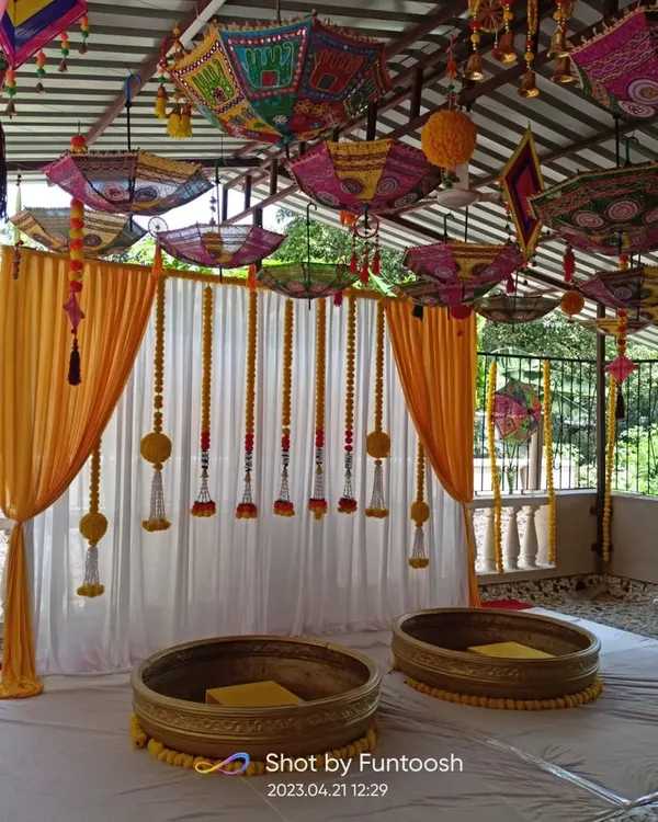 A classic Haldi ceremony setup with a canopy of colorful Rajasthani umbrellas. The stage has a simple white and yellow drape backdrop with marigold tassels.