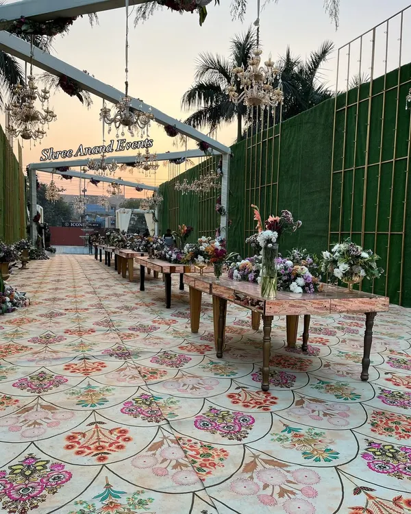 An elegant outdoor dining setup for a daytime event, featuring long wooden tables, floral arrangements, and chandeliers hanging from a white pergola.