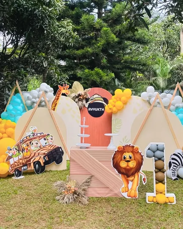 A full view of an outdoor safari camp theme for a first birthday. The setup includes teepees, a variety of animal cutouts like a lion and zebra, and balloon arrangements in earthy tones.