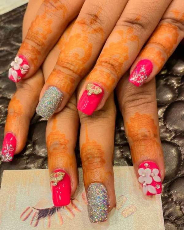 A vibrant and festive set perfect for a bride. This design combines hot pink nails, silver glitter accents, and nails with 3D sculpted flowers and bows, all complementing the henna-stained hands.