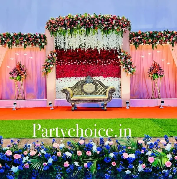 This is a grand outdoor reception stage we built. The design features a multi-layered backdrop with pink drapes, a central wall of red and white flowers, and extensive floral borders, creating a lavish and picturesque setting.