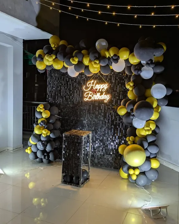 A full view of a black and yellow balloon arch framing a sparkling black sequin wall on a terrace. The setup includes a mirrored pedestal for the cake.