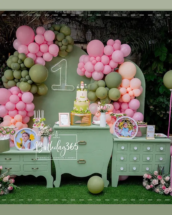 A chic and modern first birthday setup with a sage green and pink color scheme. The decor features a stylish dessert table, organic balloon arches, and a neon '1' sign, creating a fresh and beautiful photo corner.