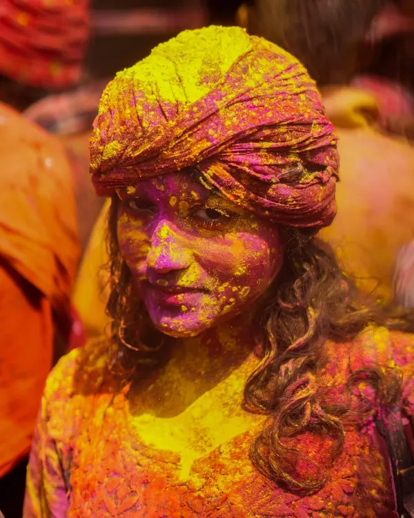 A stunning portrait of a woman at the Nandgaon Holi celebration, her face beautifully covered in yellow and pink gulal. Her direct gaze captures the joyful and immersive spirit of the festival.