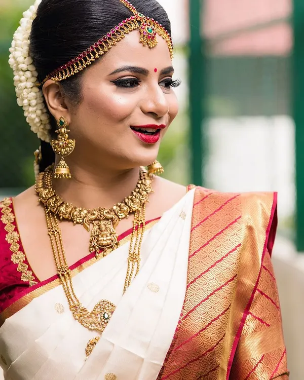 A classic South Indian bridal portrait. The look features sharp winged eyeliner, a bright red lip, and a neat hair bun adorned with fresh jasmine flowers, perfectly complementing her traditional silk saree and gold jewellery.