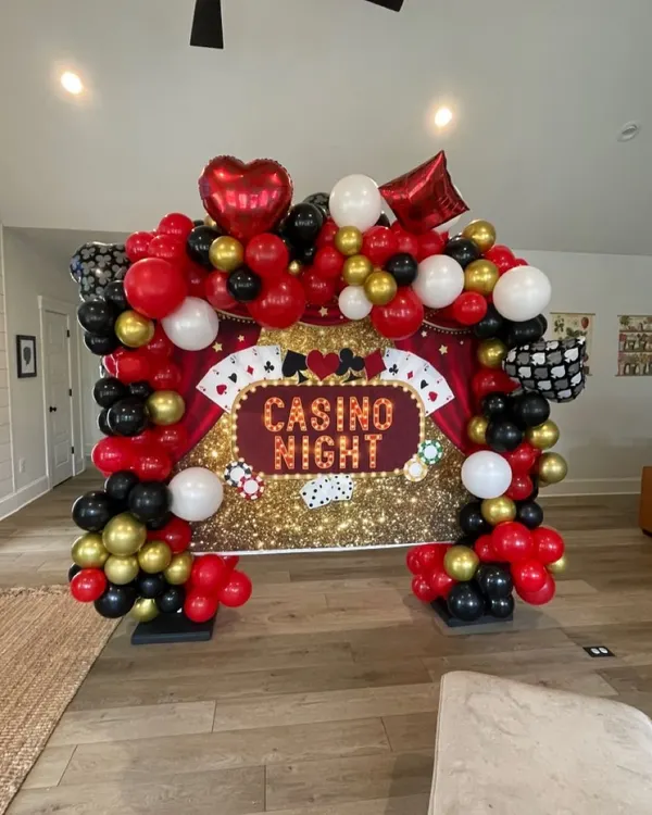A 'Casino Night' themed party backdrop. This setup features a sequined gold curtain, a balloon arch in red, black, and gold, and themed foil balloons like hearts, diamonds, and playing cards.