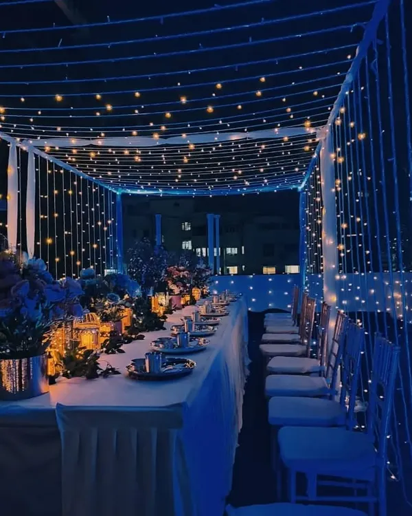 A magical rooftop dinner setup under a canopy of fairy lights. The blue ambient lighting and long table setting create a lavish and dreamy atmosphere for a special occasion.