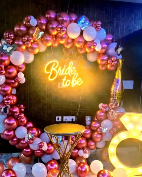This is my most popular bride-to-be party setup, featuring a rose gold and white balloon ring, a "Bride to be" neon sign, and a light-up diamond ring prop. A small stool is included for guests to sit and take photos.