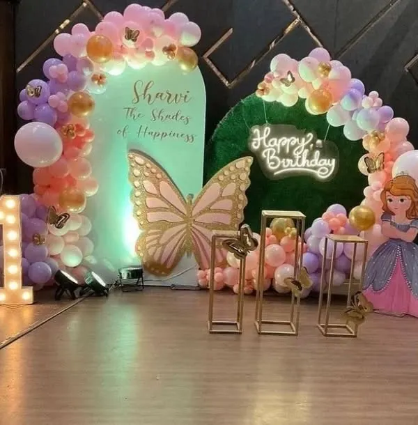 A dreamy butterfly and princess theme for Sharvi's first birthday. This elaborate setup features a large butterfly cutout, a Princess Sofia prop, pastel balloon arches, and a light-up number '1' to create a fairytale setting.