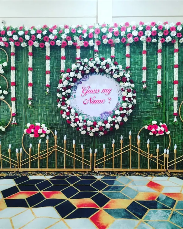 A creative name reveal idea for a naming ceremony. A floral wreath with a "Guess my Name?" sign is the centerpiece of this beautiful greenery backdrop.