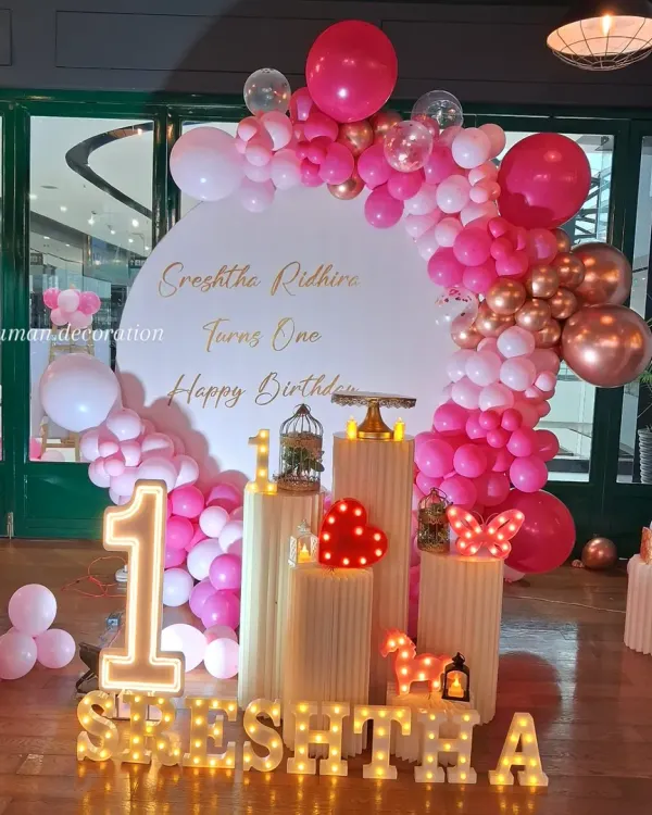 For this first birthday party, I created a full stage setup. It features a custom backdrop, a large balloon arch in shades of pink and rose gold, and multiple white plinths for the cake and props. The lighting includes a large marquee number '1' and the child's name in marquee letters, plus small decorative lights like a heart and butterfly.