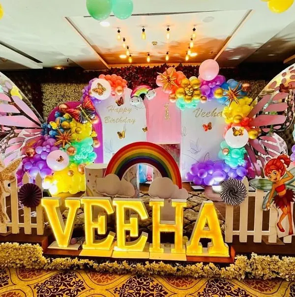 A vibrant butterfly-themed first birthday party for Veeha, featuring a colorful backdrop with large butterfly wings, rainbow accents, and light-up letters.