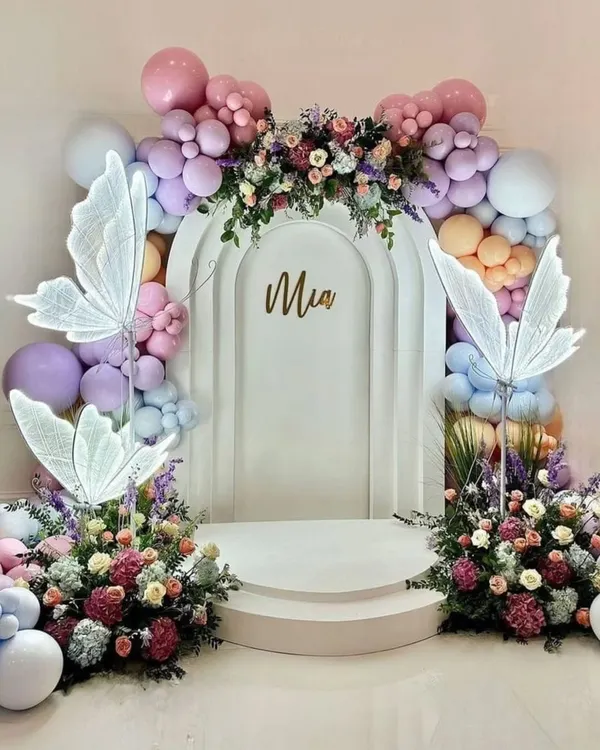 An ethereal and elegant butterfly-themed backdrop for "Mia." This design features a white arched stage, illuminated butterfly wings, and a beautiful arrangement of pastel balloons and fresh flowers, creating a dreamy photo opportunity.