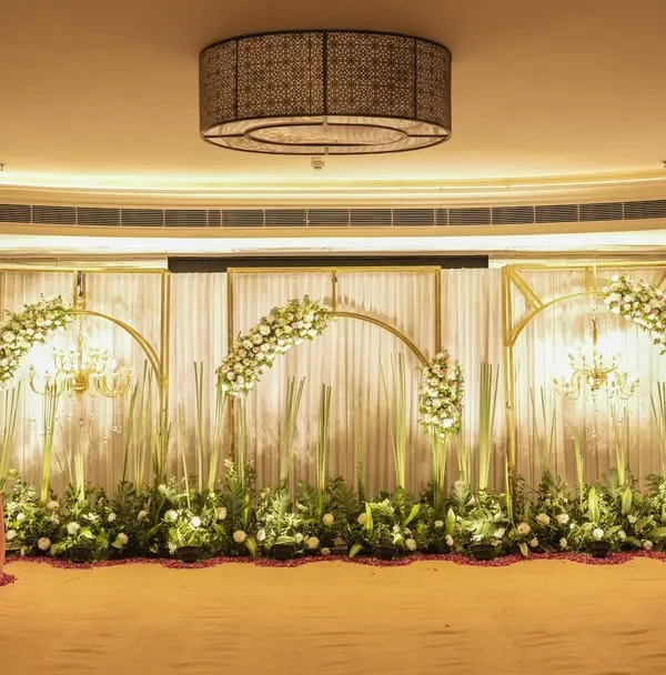 A full view of an elegant indoor stage setup, featuring golden arches, white drapes, and beautiful floral arrangements. This design creates a sophisticated and grand backdrop for a reception or engagement ceremony.