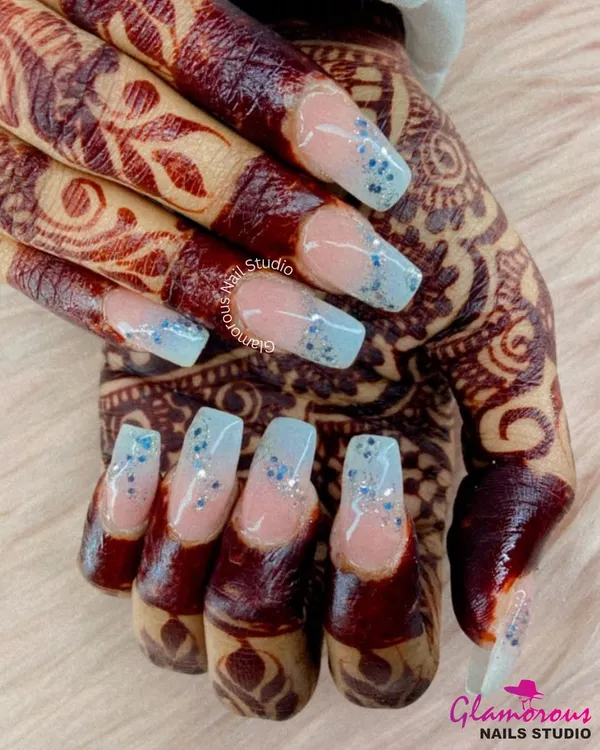 A dreamy bridal look. These square-tipped nail extensions feature a soft, natural base with a cascade of delicate blue glitter, perfectly complementing the bride's beautiful henna.