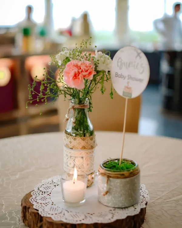 A rustic and charming table centerpiece for a baby shower. A simple arrangement of pink carnations in a lace-wrapped bottle sits on a wood slice next to a small candle, creating a warm, cozy feel.