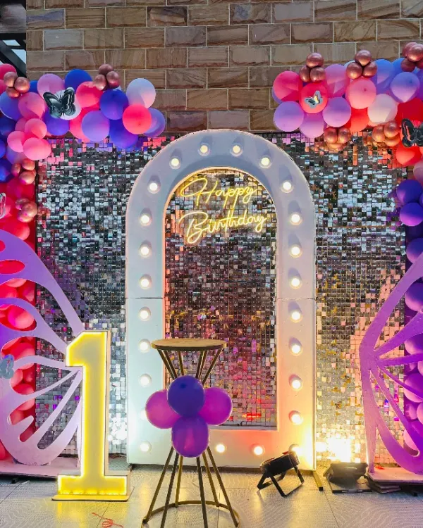 A stunning first birthday setup for an outdoor party. This features a silver shimmer wall, a large lighted marquee number '1', butterfly cutouts, and a vibrant balloon arch in pink, purple, and blue, all topped with a 'Happy Birthday' neon sign.