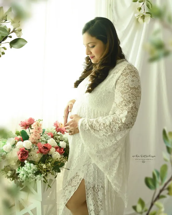 A serene moment by the window. The soft light, delicate white lace gown, and floral props create a dreamy and romantic boho-chic portrait.
