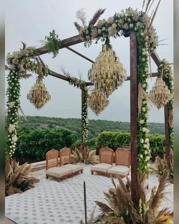 A modern rustic mandap that blends nature with luxury. This outdoor wedding setup features a wooden structure adorned with white roses and greenery, with elegant crystal chandeliers hanging above, all set against a beautiful natural backdrop.