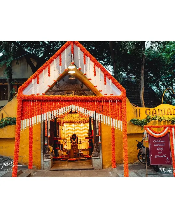 The entrance to a traditional naming ceremony at Ganjam Mantapa. The archway is heavily decorated with orange and white floral garlands, creating a grand and auspicious welcome.