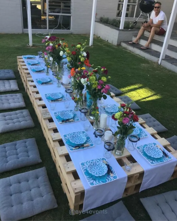 An outdoor picnic style party setup on a lush green lawn. A long, low table made from wooden pallets is set with a white runner, colorful floral arrangements, and bright blue patterned plates, with grey cushions for seating.